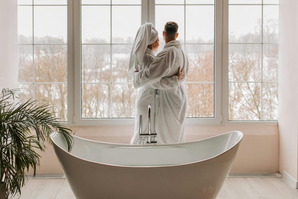 Couple in bathrobes standing beside a freestanding bathtub in a bright room with large windows and winter scenery